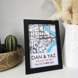 Può includere: Una stampa incorniciata con i nomi "DAN & YAZ" e la data "31 ottobre 2021". La mappa presenta un cuore rosso e sezioni blu. Accanto al quadro c'è una brocca bianca con un cuore.