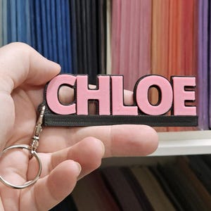 以下が含まれることがあります： 「CHLOE」という名前が書かれたピンクと黒のキーホルダー。ピンク色の文字は黒いベースに取り付けられています。シルバー色のキーリングと留め金が付いています。背景には、さまざまな色の紙があります。