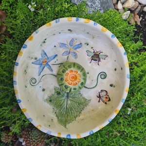 Könnte beinhalten: Eine runde Keramikschale mit einer zentralen grünen, texturierten Blütenknospe, handgemalten blauen sternförmigen Blumen, einer Hummel und einem Schmetterling. Der Rand ist mit abwechselnden gelben und blauen Strichen auf cremefarbenem Grund verziert.