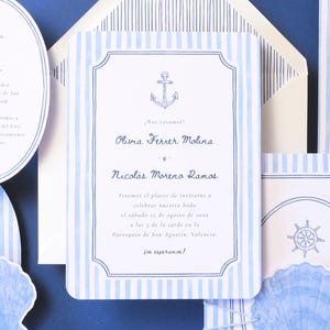 Può includere: Invito di nozze a tema nautico con carta a righe blu e bianche. L'invito include una grafica di ancora e testo, con dettagli aggiuntivi su una scheda separata. Un ritaglio a forma di conchiglia aggiunge un tocco decorativo.