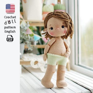 Puede incluir: Una muñeca tejida a mano con piel color café claro, cabello castaño corto y ondulado, y mejillas sonrosadas. La muñeca viste pantalones cortos de color verde claro y tiene ojos oscuros. El texto a la izquierda dice: Crochet doll pattern English PDF.