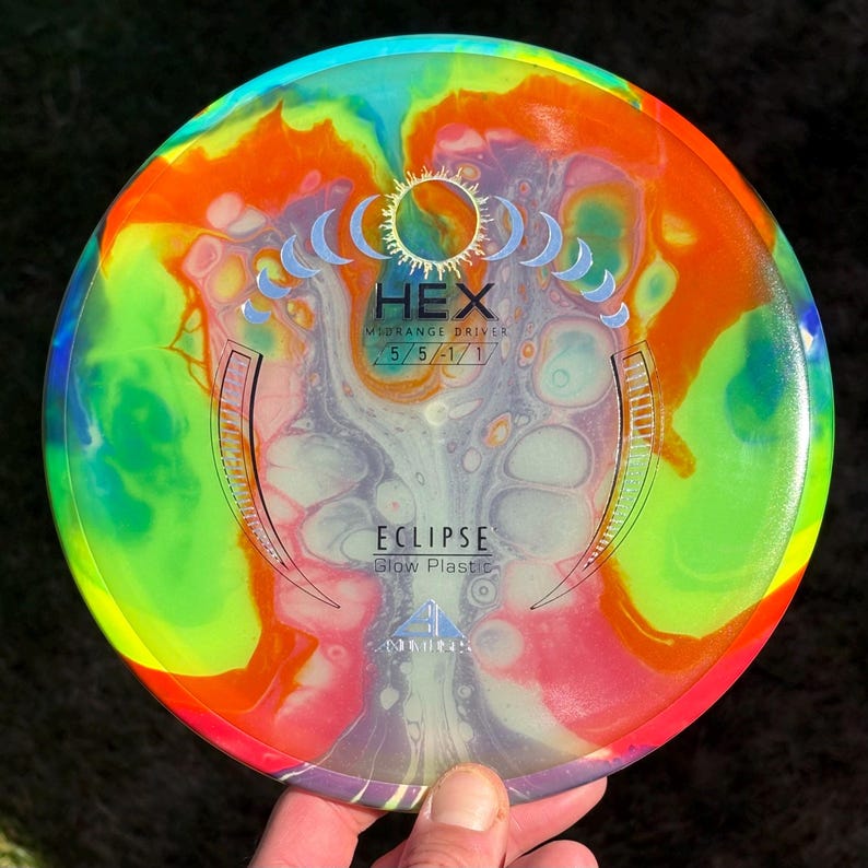 May include: A round disc golf disc featuring a vibrant, multi-coloured swirl pattern of orange, yellow, green, and purple. The centre displays the text HEX, MIDRANGE DRIVER, 5/5/-1/1, ECLIPSE, and Glow Plastic, along with a moon phase graphic.