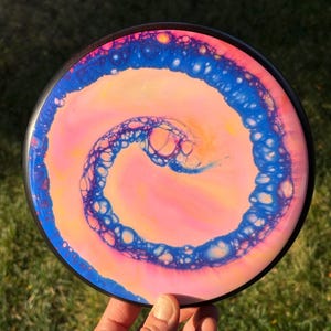 May include: A disc golf disc with a striking, swirled design. The disc has a black rim and a central pattern of pink, blue, and yellow colours. The swirling pattern resembles a galaxy or abstract artwork, making it visually appealing.