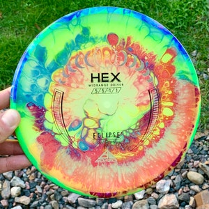 May include: A round disc golf disc featuring a vibrant, multi-coloured fluid art pattern in shades of neon yellow, orange, blue, and purple. The centre displays the text HEX, MIDRANGE DRIVER, 5/5/-1/1, ECLIPSE, Glow Plastic, and AXIOM DISCS.
