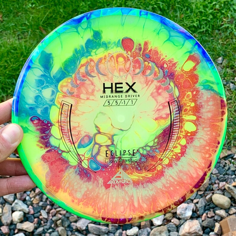 May include: A round disc golf disc featuring a vibrant, multi-coloured fluid art pattern in shades of neon yellow, orange, blue, and purple. The centre displays the text HEX, MIDRANGE DRIVER, 5/5/-1/1, ECLIPSE, Glow Plastic, and AXIOM DISCS.