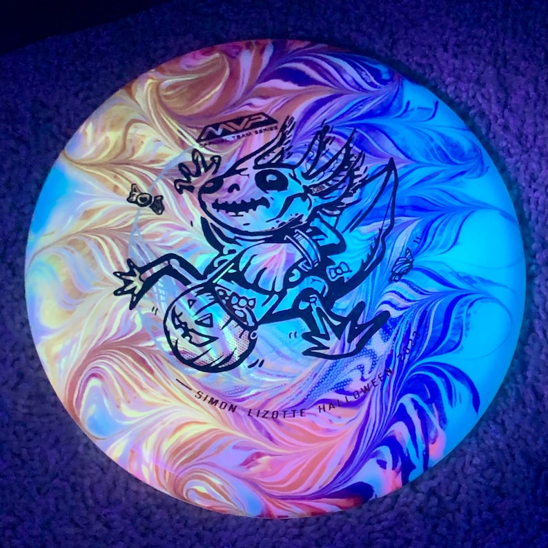 May include: A round disc golf disc featuring a swirling tie-dye pattern in shades of blue, purple, and orange. The centre displays a black graphic of a skeletal axolotl holding a jack-o'-lantern bucket, with the text MVP and SIMON LIZOTTE HALLOWEEN 2023.