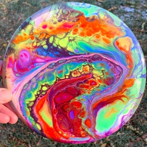 May include: A circular disc featuring a vibrant fluid acrylic pour design with swirling patterns of neon orange, bright yellow, electric blue, magenta, and lime green. The colours blend in organic, marbled shapes across the entire surface.