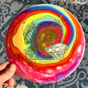 May include: A round disc golf driver with a vibrant, multi-colored swirl pattern in shades of pink, orange, yellow, blue, and purple. The center features a green graphic of a bird and a diamond, with the text DIAMOND, EASY TO USE DRIVER, 8 6 -3 1.