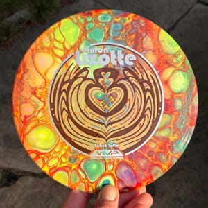 Op de afbeelding: Een ronde discgolfschijf met een levendig, meerkleurig vloeiend verfpatroon in tinten geel, oranje, rood en groenblauw. Het midden toont een bruine afbeelding van een hartvormig latte-art-ontwerp met de tekst Simon Lizotte en Bokeh Latte.