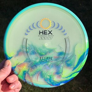May include: A round disc golf disc with a teal rim and a translucent centre featuring a swirling, multi-coloured marbled pattern of blue, green, and yellow. The centre displays the text HEX, MIDRANGE DRIVER, 5/5/-1/1, ECLIPSE, and Glow Plastic.