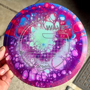 Puede incluir: Un disco de golf con un diseño abstracto y vibrante en tonos de azul, rosa, morado y verde. El disco presenta la palabra "WAVE" y los números "5 -2 2". Es un driver de distancia.