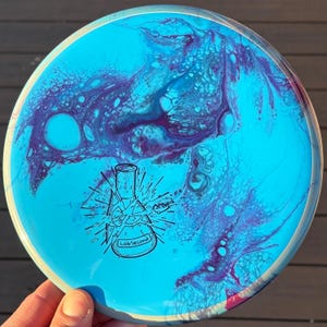 May include: A circular disc with a bright cyan base featuring a swirling purple fluid paint pattern. A black line art graphic of a shattered laboratory flask is printed in the centre with the text LabSecond written inside the flask base.