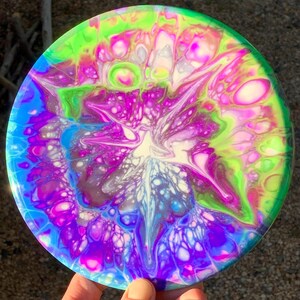 May include: A circular disc featuring a vibrant fluid acrylic pour design with swirling patterns of neon green, bright magenta, deep purple, and electric blue. The colours blend into organic, cell-like shapes across the smooth, glossy surface.