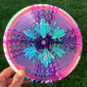 May include: A purple, pink and white disc golf disc with a swirling pattern. The disc has a holographic design of crystals in the centre with the text "OTB OPEN MMXIX" printed on the disc.