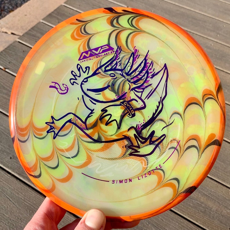 May include: A round disc golf disc featuring a swirling yellow and orange tie-dye pattern. The centre displays a purple line-art illustration of an axolotl with the text MVP, GYRO, and SIMON LIZOTTE printed in purple on the surface of the disc.