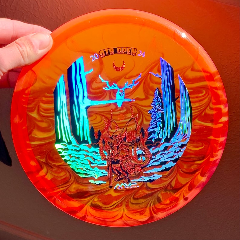 May include: A translucent orange disc golf disc featuring a holographic graphic of a forest scene with a central figure, a deer, and a crescent moon. The text 20 OTB OPEN 24 is printed at the top, and the MVP Disc Sports logo is at the bottom.