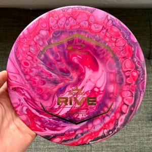 May include: A pink, purple and black disc golf disc with a gold metallic design. The disc has the words "Royal" and "Rive" in gold lettering. The disc is a "Grand" distance driver with a speed of 13, glide of 5, turn of 0, and fade of 3.5.