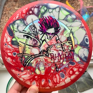 May include: Red, white, and green disc golf disc with a graphic of a drummer playing a drum set. The graphic includes the text "Rhythm Special Edition".