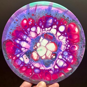 May include: A round disc with a colorful abstract design. The design features swirls of pink, purple, white, and green paint. The disc has a smooth, glossy finish.