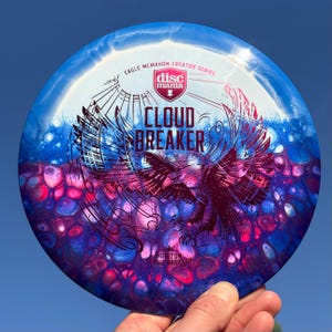 Puede incluir: Disco de golf azul y blanco con un diseño de águila y la inscripción "Cloud Breaker". El disco tiene un diseño degradado con acentos rosas y azules. También se ve el texto "Eagle McMahon Creator Series".