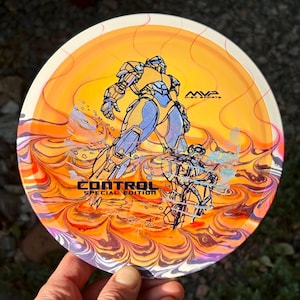 Pode incluir: Um disco de golfe redondo com um fundo marmorizado em laranja e amarelo vibrante. O gráfico retrata uma grande figura robótica em pé atrás de um piloto humano. O texto MVP DISC SPORTS e CONTROL SPECIAL EDITION está impresso em preto.