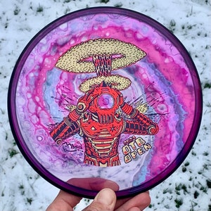 May include: A pink, white and blue disc golf disc with a red robot holding its head as a mushroom cloud explodes above it. The disc has the text "2023 OTB OPEN" printed on it.