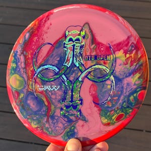 May include: A red, yellow, blue and green disc golf disc with a holographic image of a mammoth with the text "OTB OPEN" and "MVP DISC SPORTS" printed on the disc.
