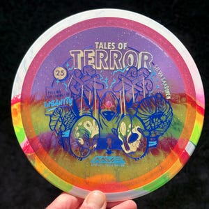 May include: A colorful disc golf disc with a graphic design. The disc features the text "TALES OF TERROR" and "PULLING THE STRINGS OF INSANITY". The disc has a purple center with a pink and white rim and a rainbow edge.