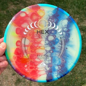 May include: A round disc golf disc featuring a vibrant fluid art pattern with red, orange, and yellow on the left and blue and purple on the right. The centre displays a moon phase graphic, the text HEX, MIDRANGE DRIVER, 5/5/-1/1, ECLIPSE, Glow Plastic, and the Axiom Discs logo.