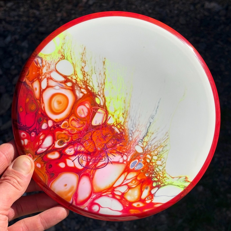 May include: A round disc with a red rim and a white centre featuring a vibrant fluid acrylic pour design in shades of orange, red, yellow, and pink. A purple line drawing of a beaker and a flask is centred on the colourful paint pattern.