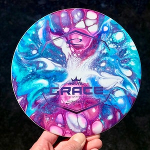 May include: A disc golf disc with a vibrant, marbled pattern in blue, purple, and white. The disc displays the text "ROYAL GRACE" and "GRAND DISTANCE DRIVER". The disc is held by a hand.