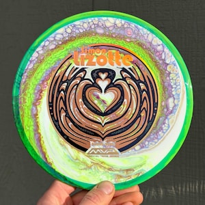 May include: A round disc golf disc featuring a vibrant fluid art pattern in shades of neon green, purple, and white. The center displays a black graphic design with heart-like shapes and the text Simon Lizotte, MVP, and Official Team Series.