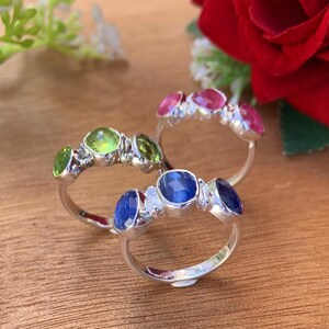 May include: Three silver rings with colourful gemstones. The first ring has three green gemstones, the second ring has three red gemstones, and the third ring has three blue gemstones with a small diamond between each.