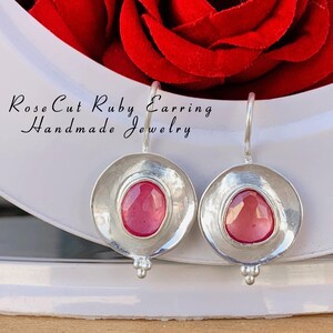 Puede incluir: Un par de pendientes de plata con piedras preciosas de rubí talla rosa. Los pendientes están engastados en un engaste redondo de plata martillada. Los pendientes cuelgan de un gancho de plata.