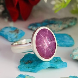 Op de afbeelding: Een zilveren ring met een roze ster-robijn edelsteen. De ring ligt op een bed van turquoise stenen.