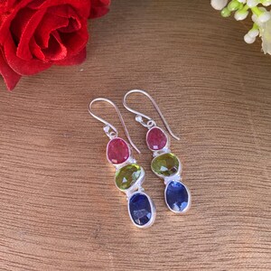 May include: A pair of silver dangle earrings with three gemstone drops. The top drop is red, the middle is green, and the bottom is blue.