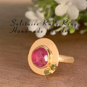 May include: A gold ring with a large pink ruby gemstone and two smaller green gemstones. The ring is set in a gold band with a raised oval setting. The text "Solitaire Ruby Ring Handmade Jewelry" is visible in the image.