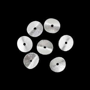 Może przedstawiać: Siedem srebrnych koralików w kształcie dysku, z radialnym wzorem tekstury i centralnym otworem. Koraliki ułożone są na czarnym tle, eksponując ich metaliczny połysk i design. Idealne do tworzenia biżuterii lub projektów rzemieślniczych.