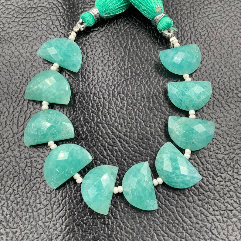 Puede incluir: Un collar de cuentas de piedra preciosa Amazonita. Las cuentas son de color verde azulado claro, cortadas en forma de media luna y facetadas. El collar est&aacute; ensartado en un cord&oacute;n blanco y terminado con una borla verde.