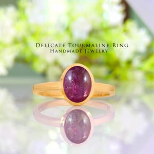 Op de afbeelding: Een gouden ring met een ovale, dieppaarse toermalijn edelsteen. De ring is gezet in een gouden band en weerspiegelt op een spiegelend oppervlak. De tekst "DELICATE TOURMALINE RING HANDMADE JEWELRY" staat boven de ring.