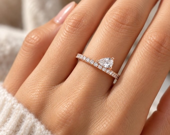 May include: A rose gold ring featuring a single pear-shaped clear stone set at an angle on a band encrusted with a row of small, round, clear pavé stones. The band has a polished metallic finish and follows a straight line across the finger.