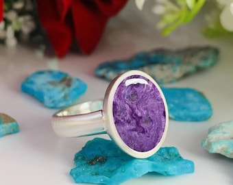 May include: A silver ring featuring an oval, purple gemstone with a swirling pattern. The stone is set in a silver bezel. The ring is displayed on a white surface with turquoise stones and flowers in the background.
