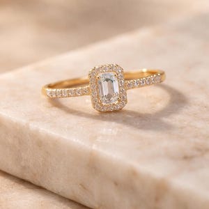 Könnte beinhalten: Ein goldener Ring mit einem rechteckigen, hellfarbenen Mittelstein, umgeben von kleineren, runden Steinen. Das Band ist ebenfalls mit kleinen Steinen verziert. Der Ring ist vor einem neutralen Hintergrund platziert.