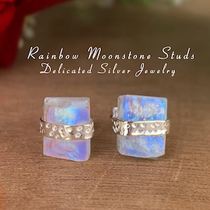 Puede incluir: Un par de pendientes de botón de piedra lunar arcoíris con una banda plateada alrededor de la piedra. La banda plateada tiene una textura martillada. Los pendientes están sobre una superficie de madera. Rainbow Moonstone Studs Delicated Silver Jewelry