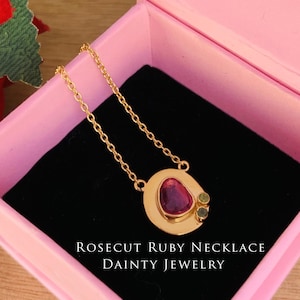 Könnte beinhalten: Eine Halskette mit einem rosenschliffenen Rubin, einer goldenen Kette und einem runden Anhänger mit einem rosafarbenen Rubin und zwei grünen Edelsteinen. Die Halskette befindet sich in einer rosa Schachtel mit dem Text "ROSECUT RUBY NECKLACE DAINTY JEWELRY".