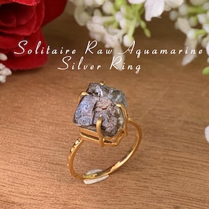 May include: A silver ring with a raw aquamarine gemstone. The ring is set in a gold band and has a simple design. The gemstone is a light blue color and has a rough, natural texture.