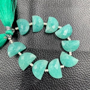 Puede incluir: Un hilo de piedras preciosas de amazonita, cada una cortada en forma de media luna con superficies facetadas. Las piedras son de color verde azulado claro y est&aacute;n ensartadas con cuentas blancas. Una borla verde azulado es visible en el lado izquierdo.