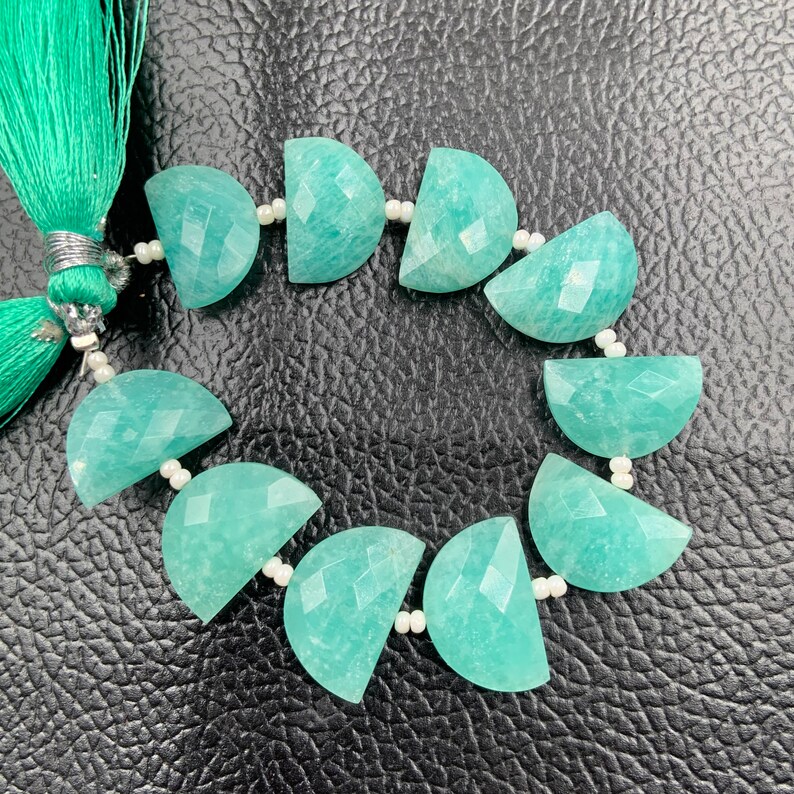 Puede incluir: Un hilo de piedras preciosas de amazonita, cada una cortada en forma de media luna con superficies facetadas. Las piedras son de color verde azulado claro y est&aacute;n ensartadas con cuentas blancas. Una borla verde azulado es visible en el lado izquierdo.