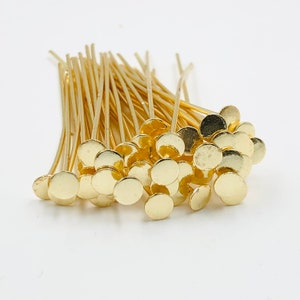 May include: A pile of gold-colored head pins with round, flat heads. The pins are arranged in a random pattern, with some of the heads overlapping.