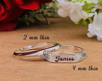 May include: Two silver rings, one thin and engraved with "Oliver", the other thicker and engraved with "James". The thin ring is 2 mm, the thicker ring is 4 mm. The rings are on a wooden surface.